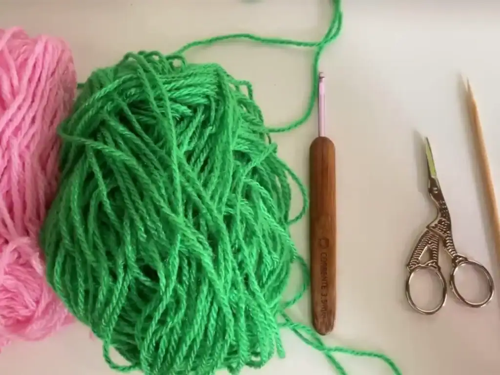 The materials needed for the crochet rose pattern: pink and green yarn, a crochet hook, scissors, and a wooden skewer.