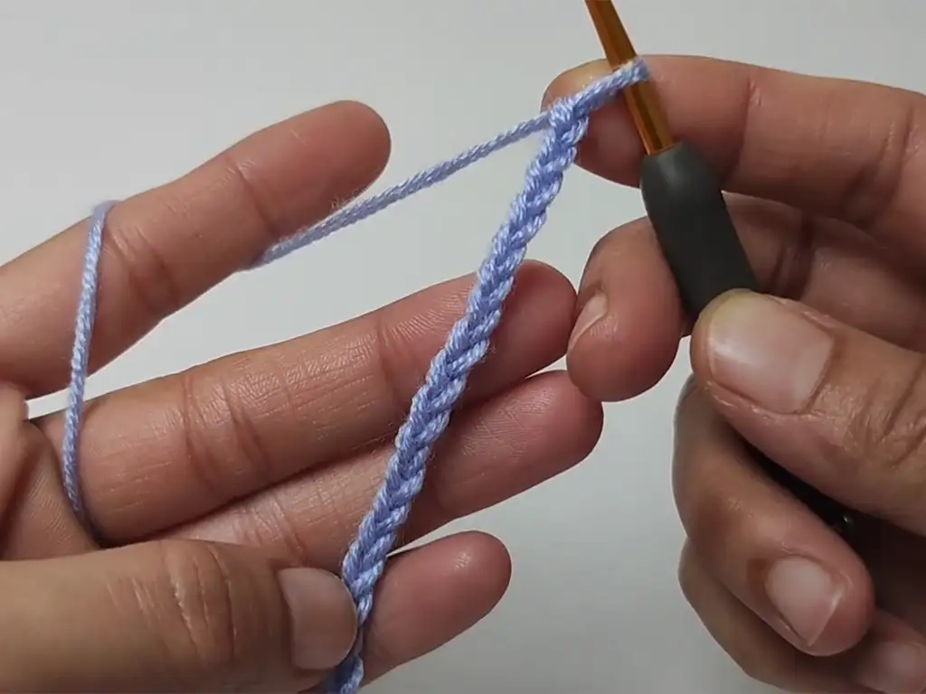 A close-up image of a crocheter's hands holding a crochet hook and working a foundation chain in pastel purple yarn.