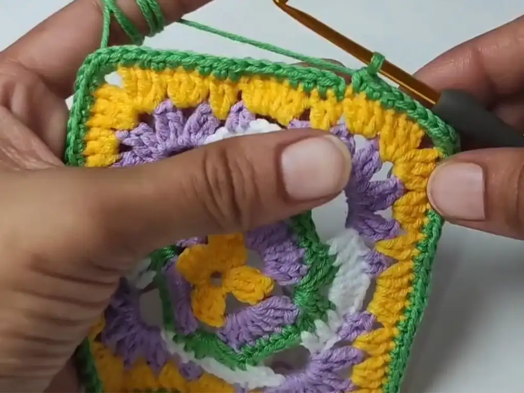 Adding the final round of stitches to a colorful crochet granny square using yellow yarn.