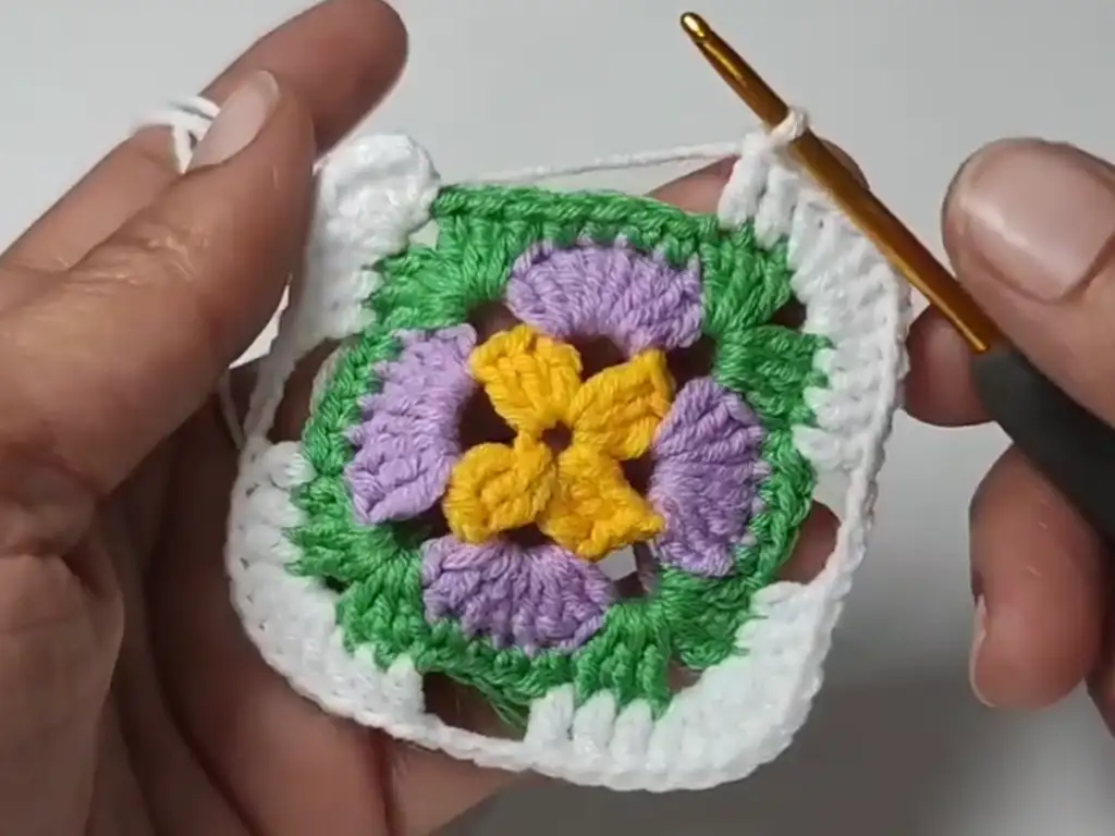 A crochet granny square with four completed rounds, showcasing a cheerful color palette of yellow, purple, green, and white.