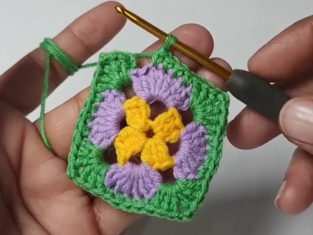 A crochet granny square after three rounds, showing a colorful pattern with yellow, purple, and green yarn.