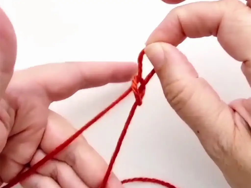 Hands demonstrating how to make a slip knot with red yarn.
