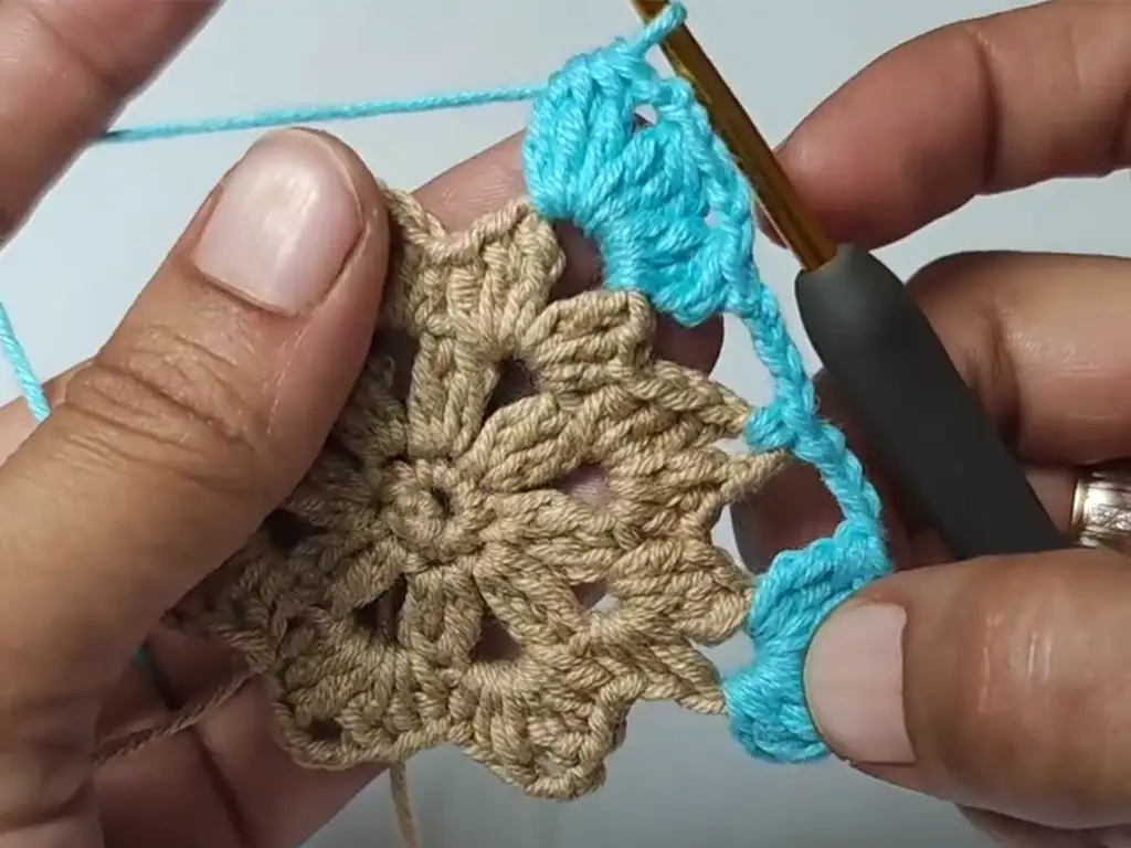 A hand holding a crochet hook, continuing to work the blue border on the crochet square pattern.