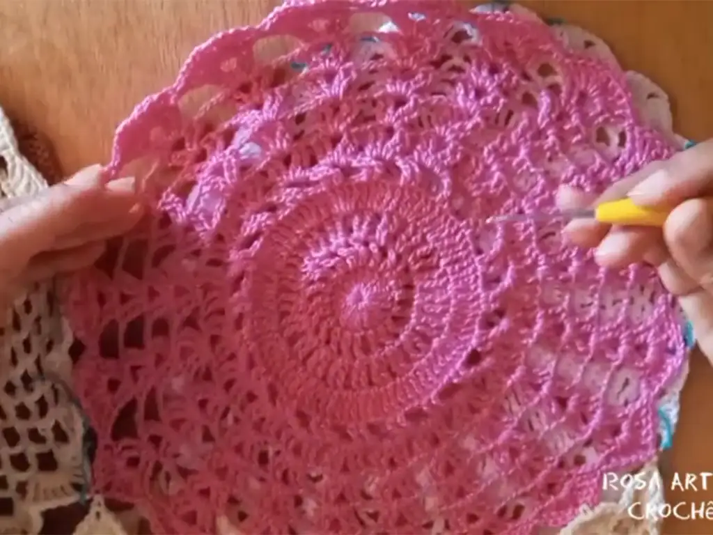 A crocheter working on a pink crochet circle motif, using a yellow crochet hook, demonstrating the creation of a dress panel.