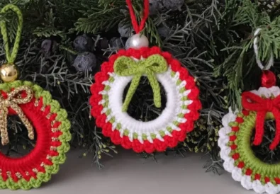 Three handmade crochet Christmas wreath ornaments in red, white, and green, hanging on a festive evergreen bough.