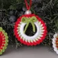 Three handmade crochet Christmas wreath ornaments in red, white, and green, hanging on a festive evergreen bough.