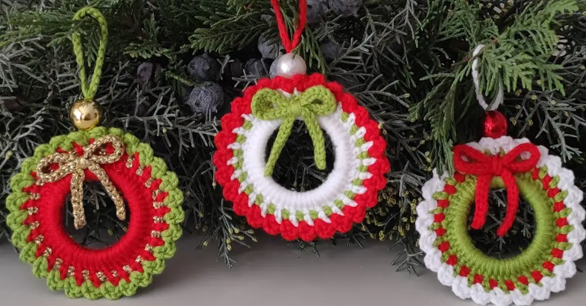 Three handmade crochet Christmas wreath ornaments in red, white, and green, hanging on a festive evergreen bough.