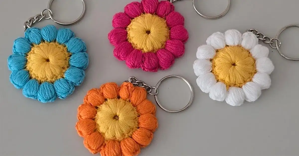 Four colorful crochet flower keychains in blue, pink, white, and orange, with yellow centers, displayed against a white background.