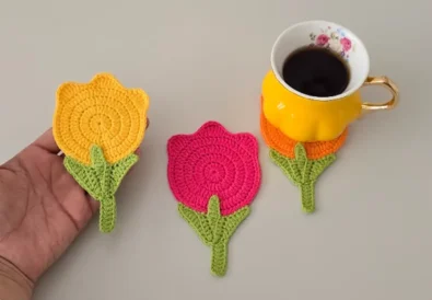 Hand holding a crochet tulip coaster in orange and pink next to coffee cups