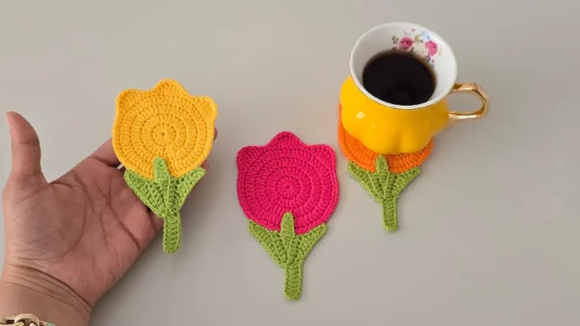 Hand holding a crochet tulip coaster in orange and pink next to coffee cups