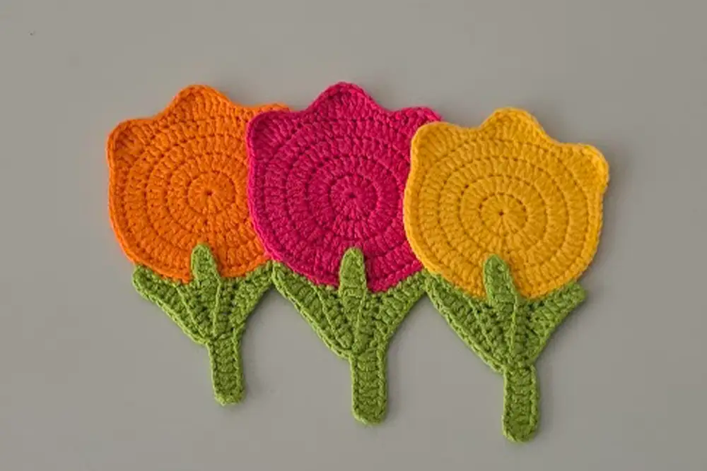 Three tulip crochet coasters in orange, pink, and yellow aligned on a table