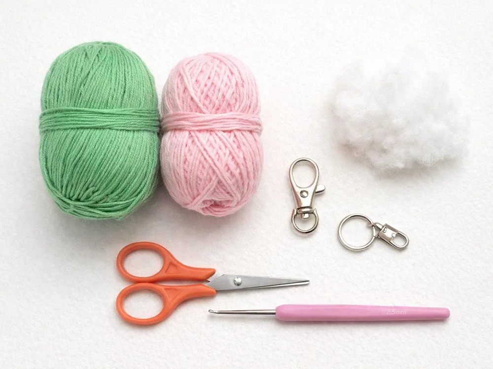 Easy crochet hat keychain materials including cotton yarn, 2.5 mm hook, scissors, keychain clip and fiberfill arranged on a white background.