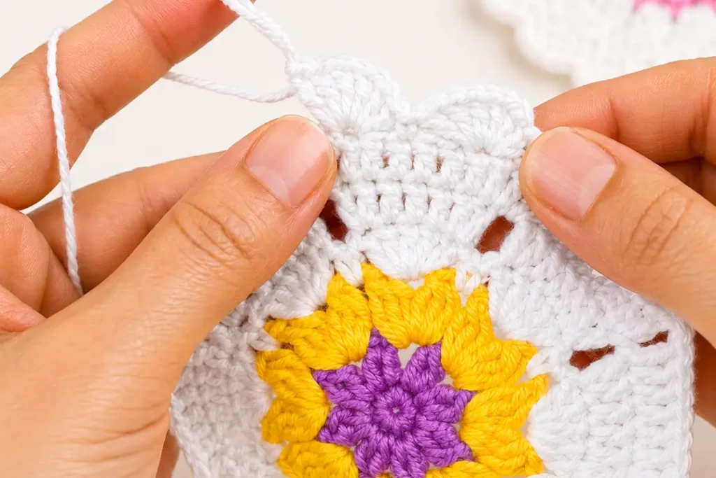 Close-up of the outer round in a crochet heart coaster pattern, showing the white stitches being worked around the yellow and purple center