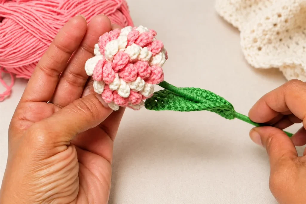 Realistic crochet flower bouquet with pink and cream petals, green crochet leaf, and matching pink cotton yarn on a clean craft table.