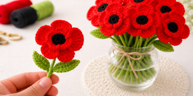 Hand holding a realistic crochet poppy flower next to a bouquet of red crochet poppies in a glass vase.
