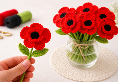 Hand holding a realistic crochet poppy flower next to a bouquet of red crochet poppies in a glass vase.