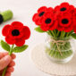 Hand holding a realistic crochet poppy flower next to a bouquet of red crochet poppies in a glass vase.