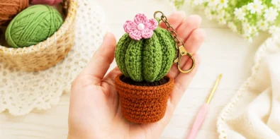 Finished crochet cactus keychain held in hand with cotton yarn balls and crochet hook on a soft pastel craft table.