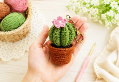 Finished crochet cactus keychain held in hand with cotton yarn balls and crochet hook on a soft pastel craft table.