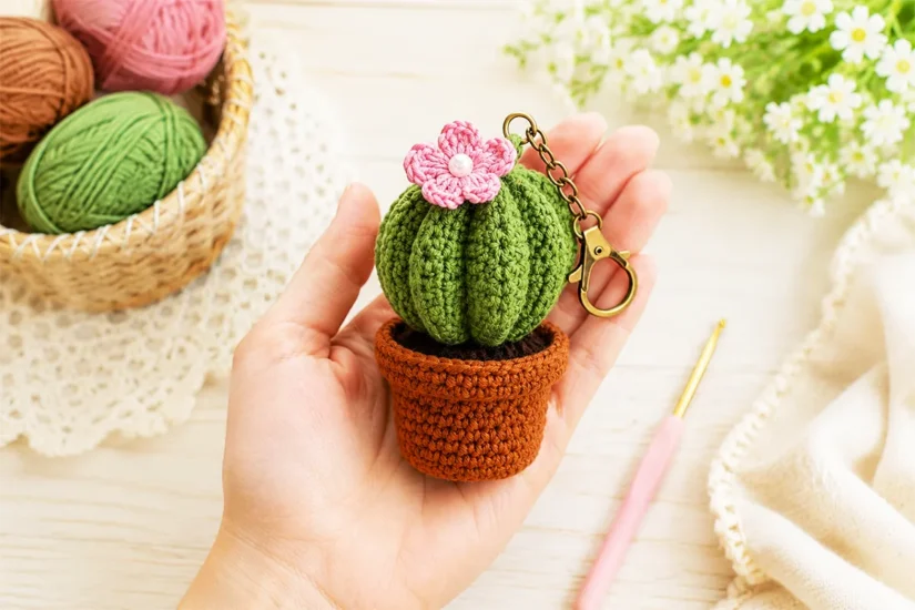 Finished crochet cactus keychain held in hand with cotton yarn balls and crochet hook on a soft pastel craft table.