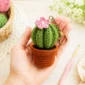 Finished crochet cactus keychain held in hand with cotton yarn balls and crochet hook on a soft pastel craft table.