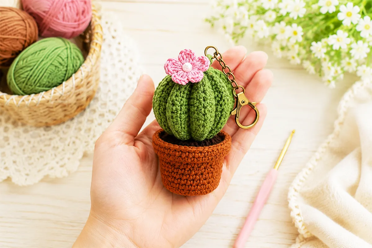 Finished crochet cactus keychain held in hand with cotton yarn balls and crochet hook on a soft pastel craft table.