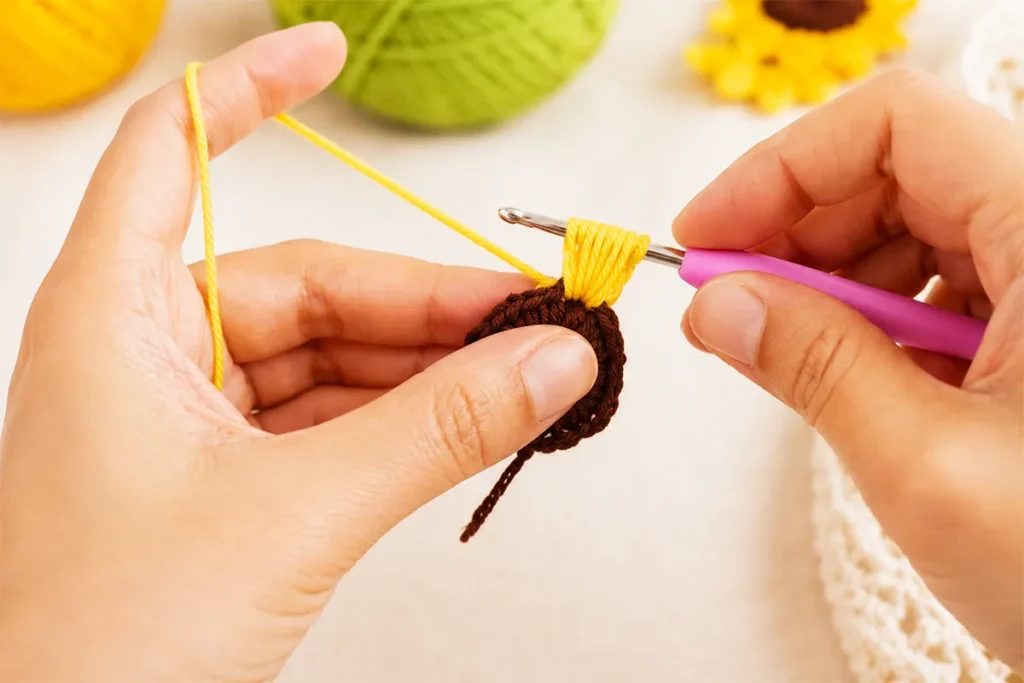 Hands adding yellow puff stitch petals to a brown crochet sunflower center with a pink crochet hook
