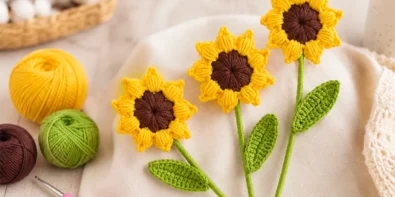 Realistic crochet sunflower with yellow puff stitch petals, brown center, green stems, and crochet leaves on a bright craft table