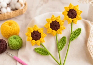 Realistic crochet sunflower with yellow puff stitch petals, brown center, green stems, and crochet leaves on a bright craft table