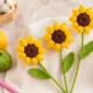 Realistic crochet sunflower with yellow puff stitch petals, brown center, green stems, and crochet leaves on a bright craft table