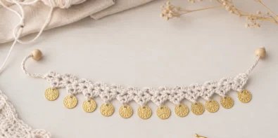 Crochet coin charm anklet made with white cotton yarn and gold coin charms on a soft neutral craft table with yarn and crochet hook
