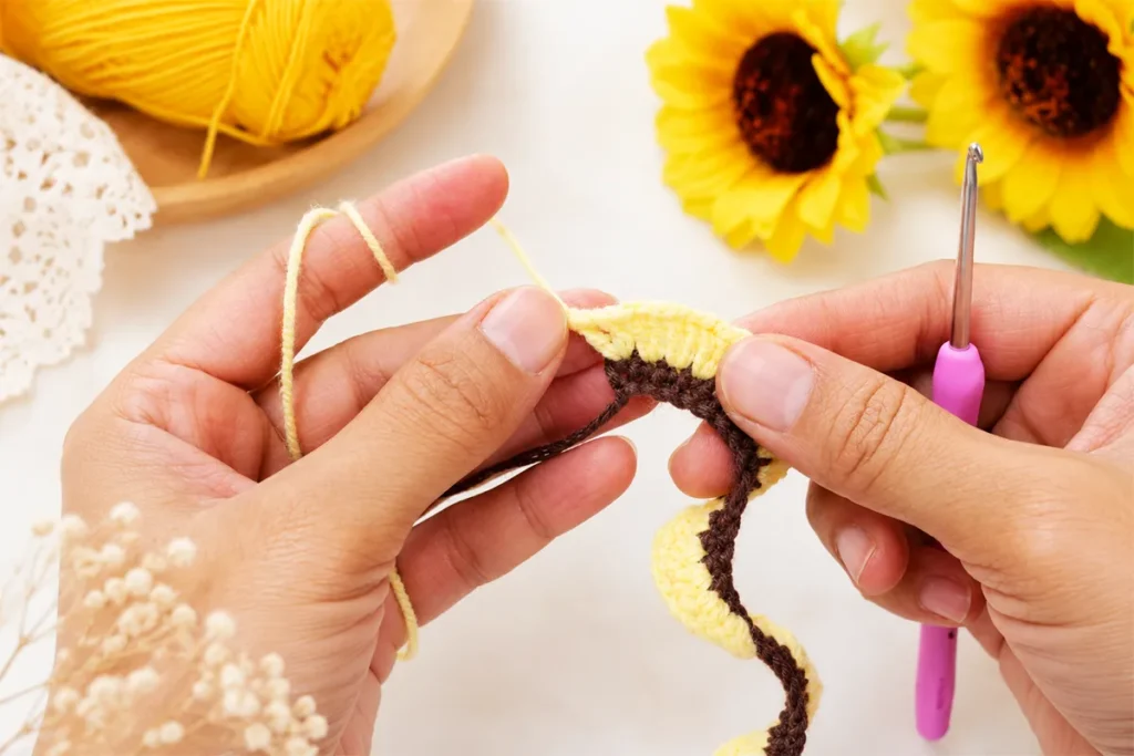 Hands making the V-stitch row with pale yellow yarn over a brown crochet base for a crochet sunflower wind spinner pattern