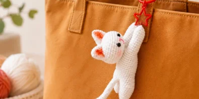 White crochet cat keychain with pink ears and a red star clasp hanging from a brown tote bag.