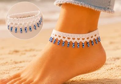 White crochet evil eye anklet with dangling blue charms displayed on a soft neutral craft table with cotton yarn and a crochet hook.