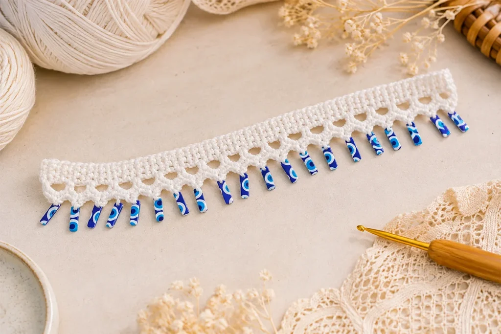 Finished white crochet evil eye anklet with dangling blue rectangular charms displayed on a neutral craft table with yarn and a crochet hook.