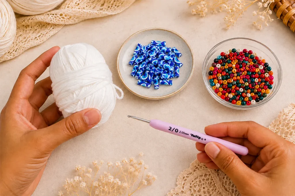Hands holding white cotton yarn and a pink 2.0 mm crochet hook beside evil eye charms and colorful beads on a neutral craft table.