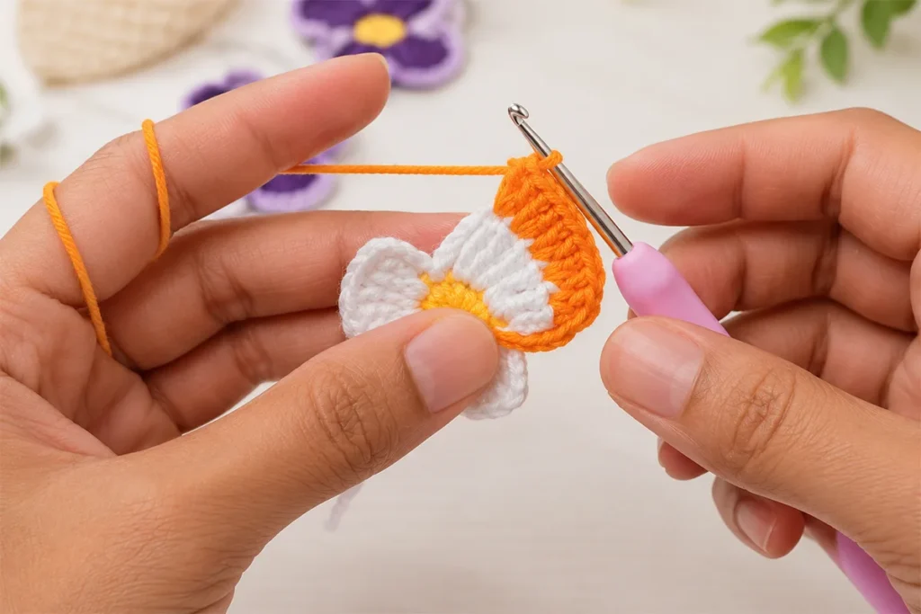 Hands crocheting the orange outer petal round around a white pansy flower motif with a crochet hook.