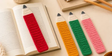 Four crochet pencil bookmarks in pink, red, yellow, and green, with one bookmark held above an open book on a warm neutral desk.