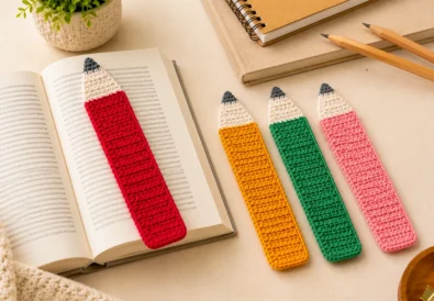 Four crochet pencil bookmarks in pink, red, yellow, and green, with one bookmark held above an open book on a warm neutral desk.