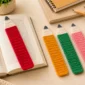 Four crochet pencil bookmarks in pink, red, yellow, and green, with one bookmark held above an open book on a warm neutral desk.