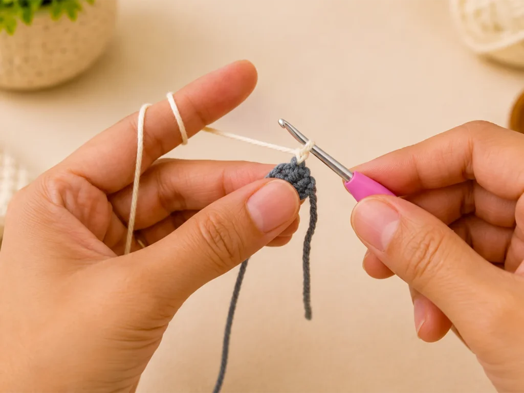 Close-up of hands crocheting a small dark gray pencil tip and changing to light cream yarn with a pink crochet hook.