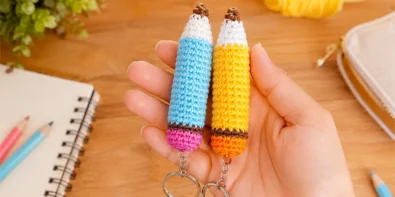 Two crochet pencil keychains in blue and yellow held in a hand with school supplies in the background