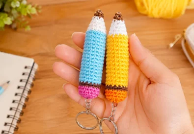 Two crochet pencil keychains in blue and yellow held in a hand with school supplies in the background
