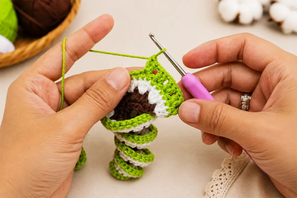 Hands holding a tree-inspired crochet wind spinner while working the green row with a pink 2.5 mm crochet hook in a close-up tutorial step.
