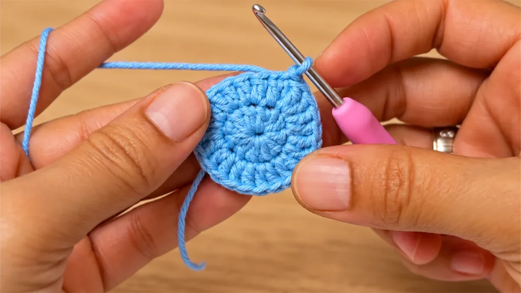Hands holding the blue crochet center of a snowflake coaster with a pink-handled crochet hook.