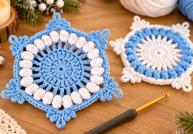 Crochet snowflake coaster with blue center, white popcorn stitches and a reversed-color coaster in the background
