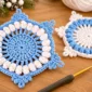 Crochet snowflake coaster with blue center, white popcorn stitches and a reversed-color coaster in the background