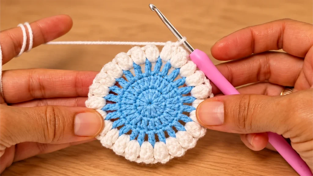 Blue crochet snowflake coaster with a ring of raised white popcorn stitches held between two hands.